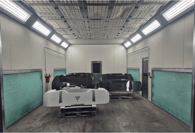 Paint booth interior with bright overhead lights and car body panels resting on pallets in the center of the bay.