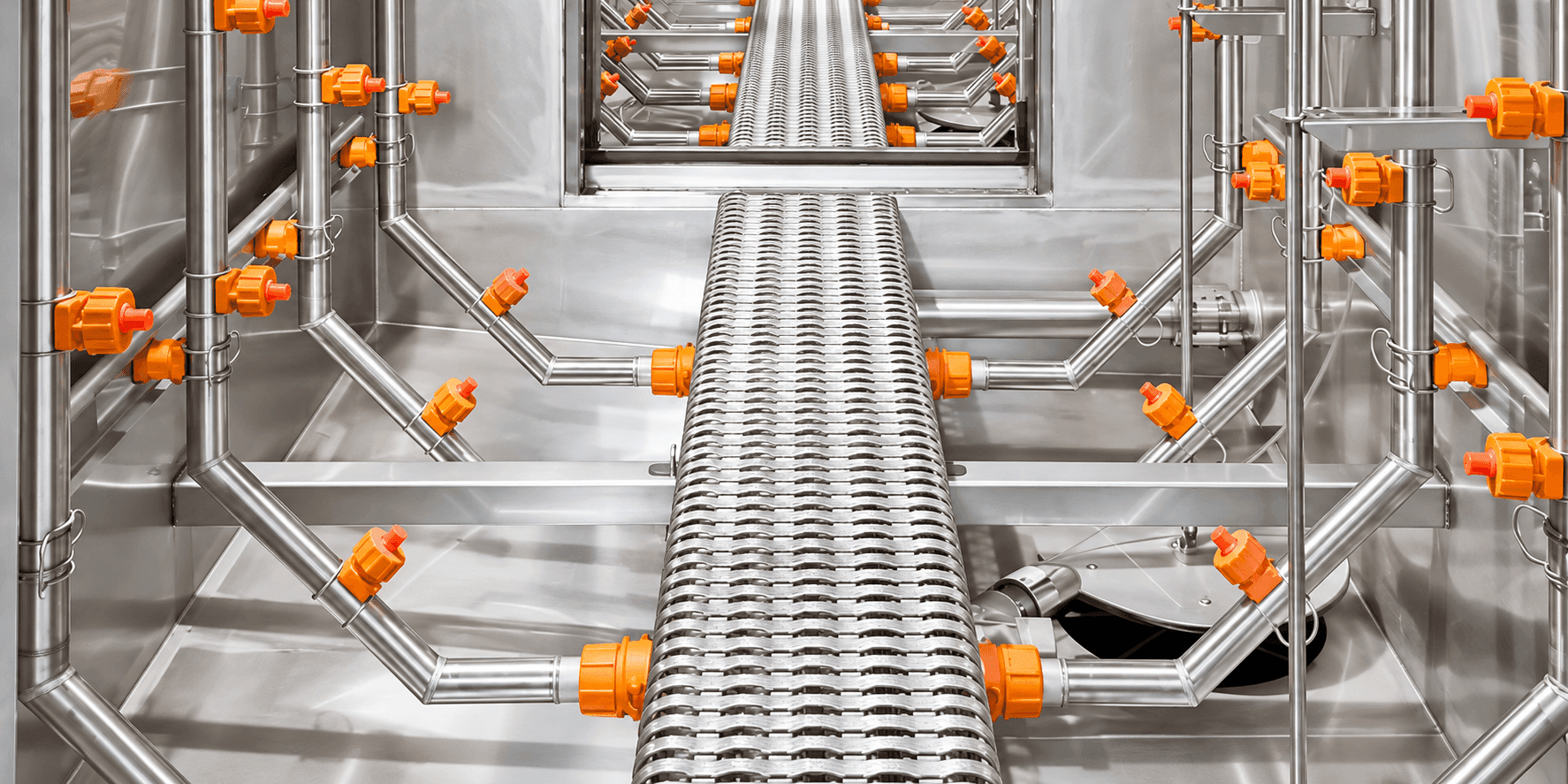 Stainless steel industrial piping system with orange valves surrounding a perforated central conveyor belt.